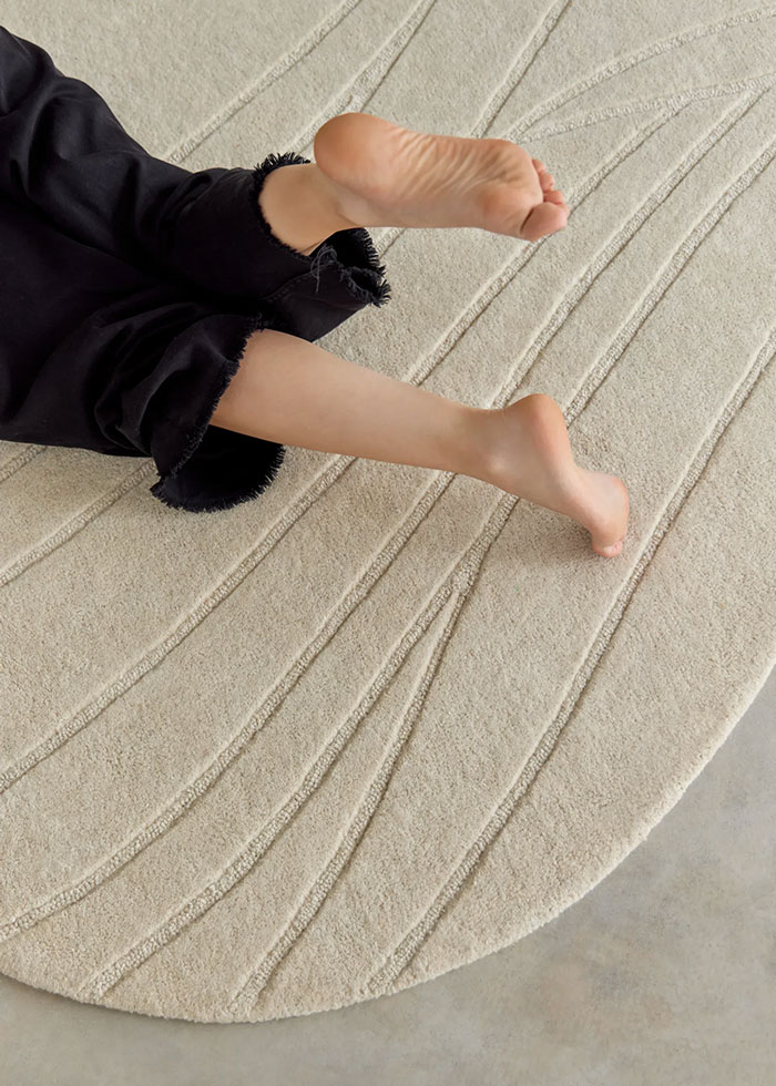 Alfombra de color hueso mullida con dibujo de líneas sinuosas y encima unas piernas de mujer con pies descalzos.