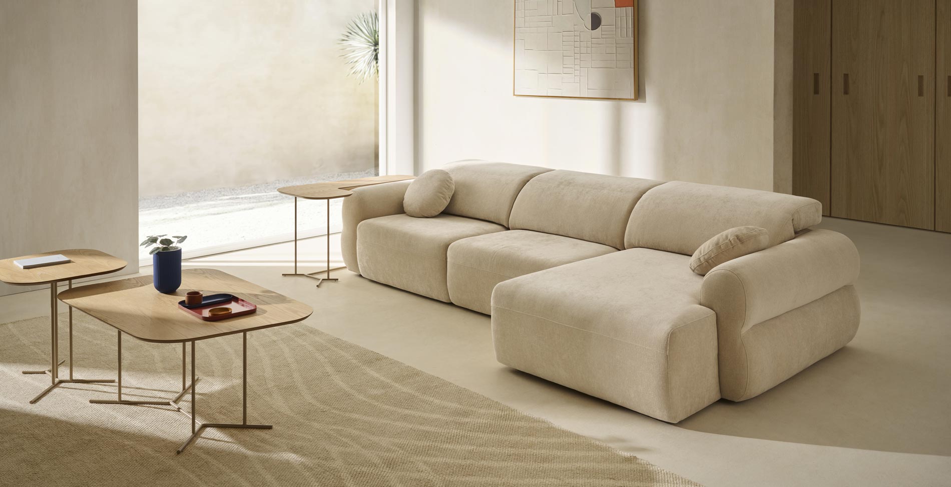 Sofá curvo con reposabrazos redondos tapizado en color crema en un salón con luz natural y tonos arena.